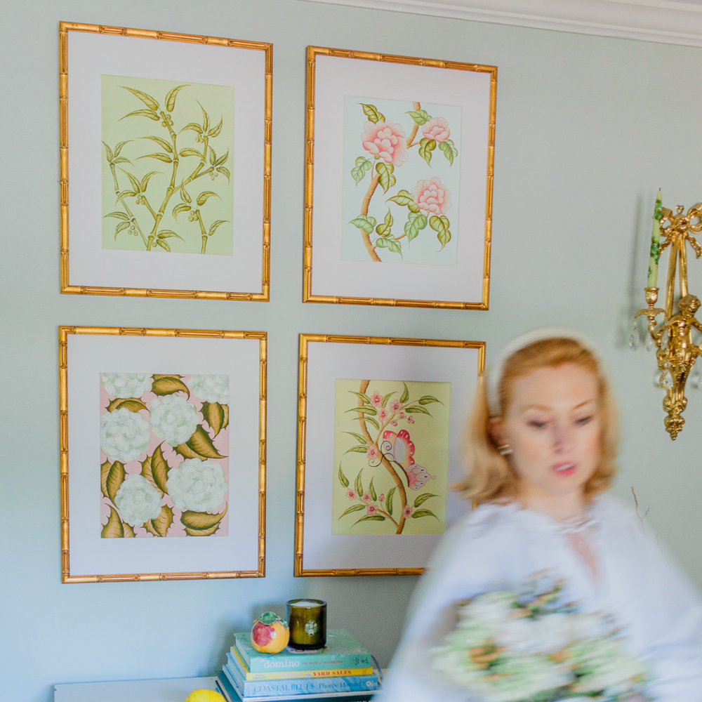 Four framed floral prints in gold bamboo frames on a pale blue wall above a small table with stacked books, a blurred woman walking by holding a bouquet