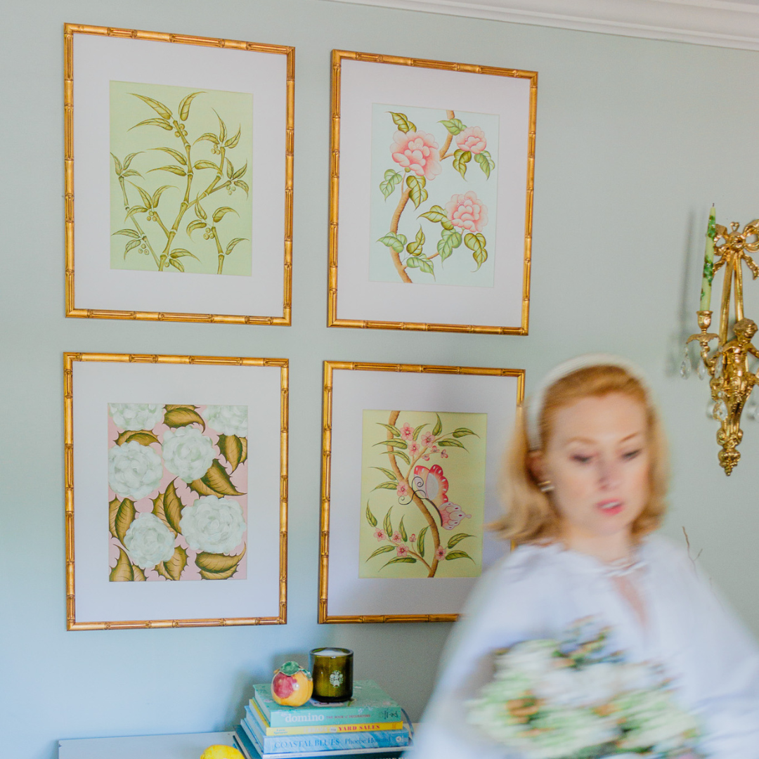 Four framed floral prints in gold bamboo frames on a pale blue wall above a small table with stacked books, a blurred woman walking by holding a bouquet