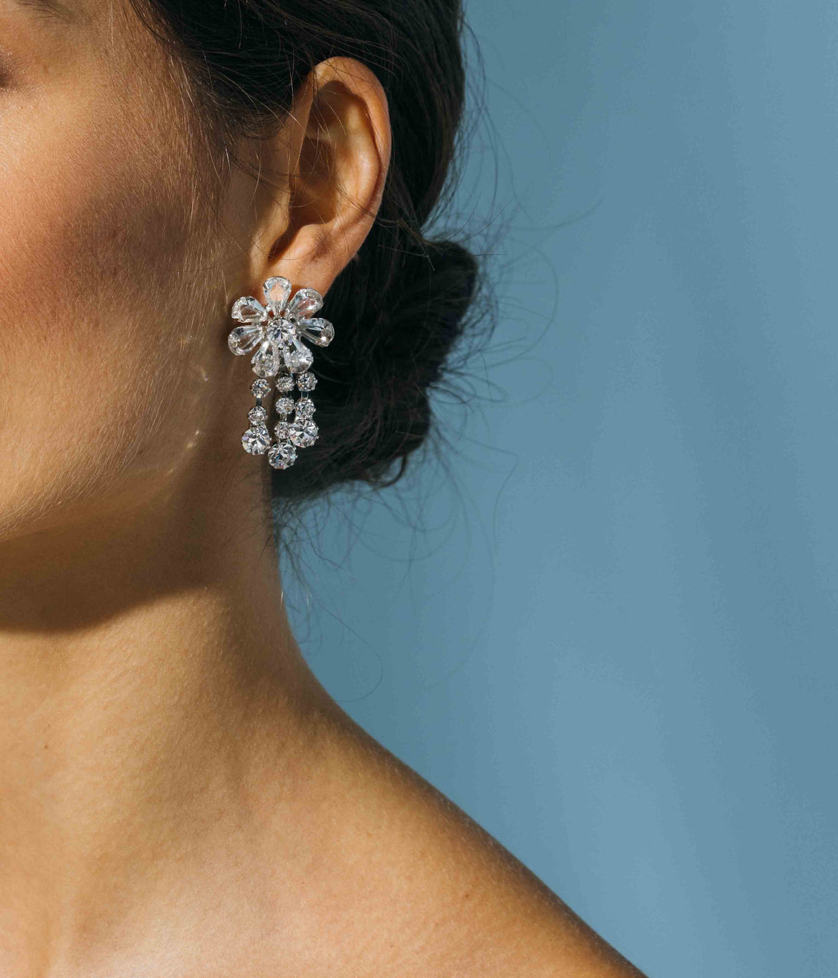 Close-up side view of Frances Earrings in Crystal showcasing a daisy flower and cascading crystal fringe on an ear against a blue background
