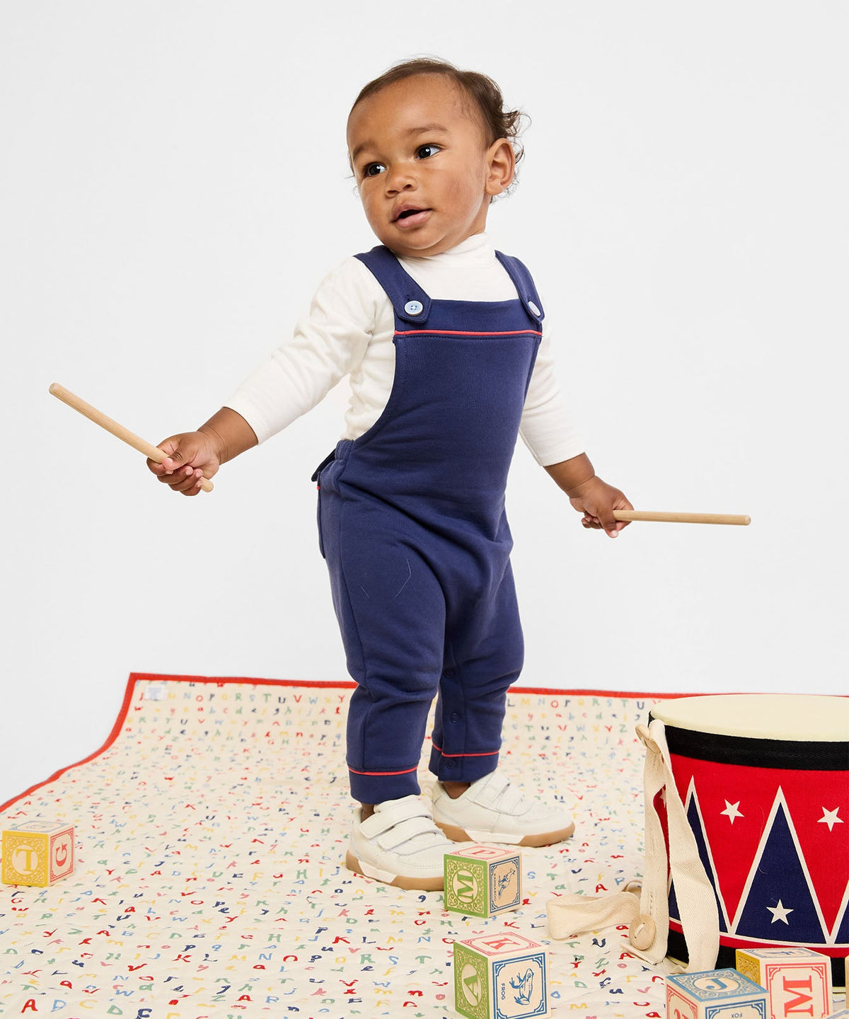 Toddler standing on alphabet play mat holding drumsticks, wearing Frank T-Neck onesie in vanilla piping layered under navy overalls