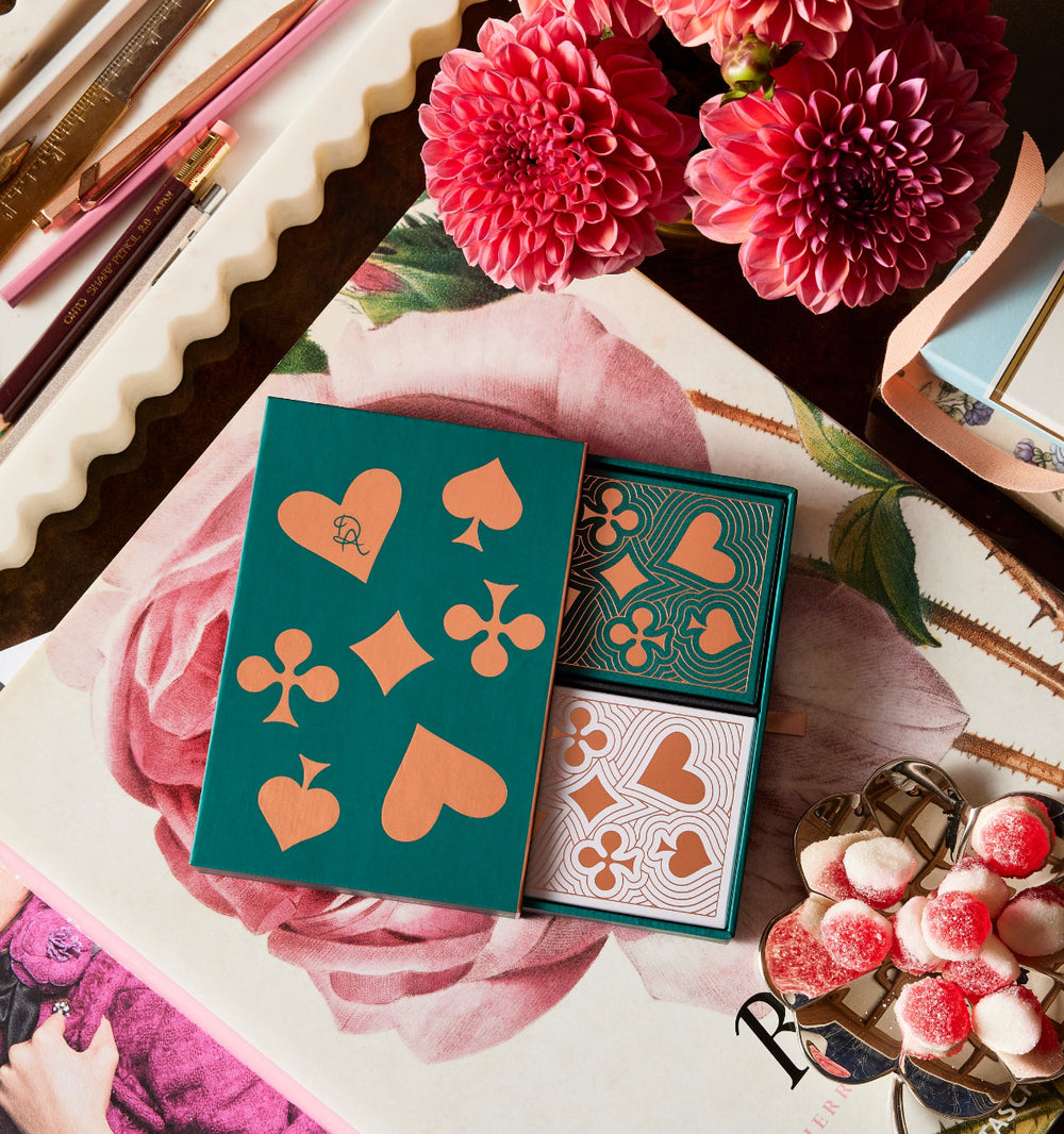 Top-down view of Lady Luck playing cards in a teal box showing copper foil suit motifs and two patterned decks on a floral tabletop