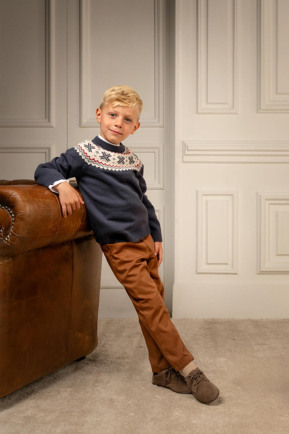 Three-quarter view of George Pants in chestnut on a child model showing tailored slim fit and ankle-length rise by a leather armchair