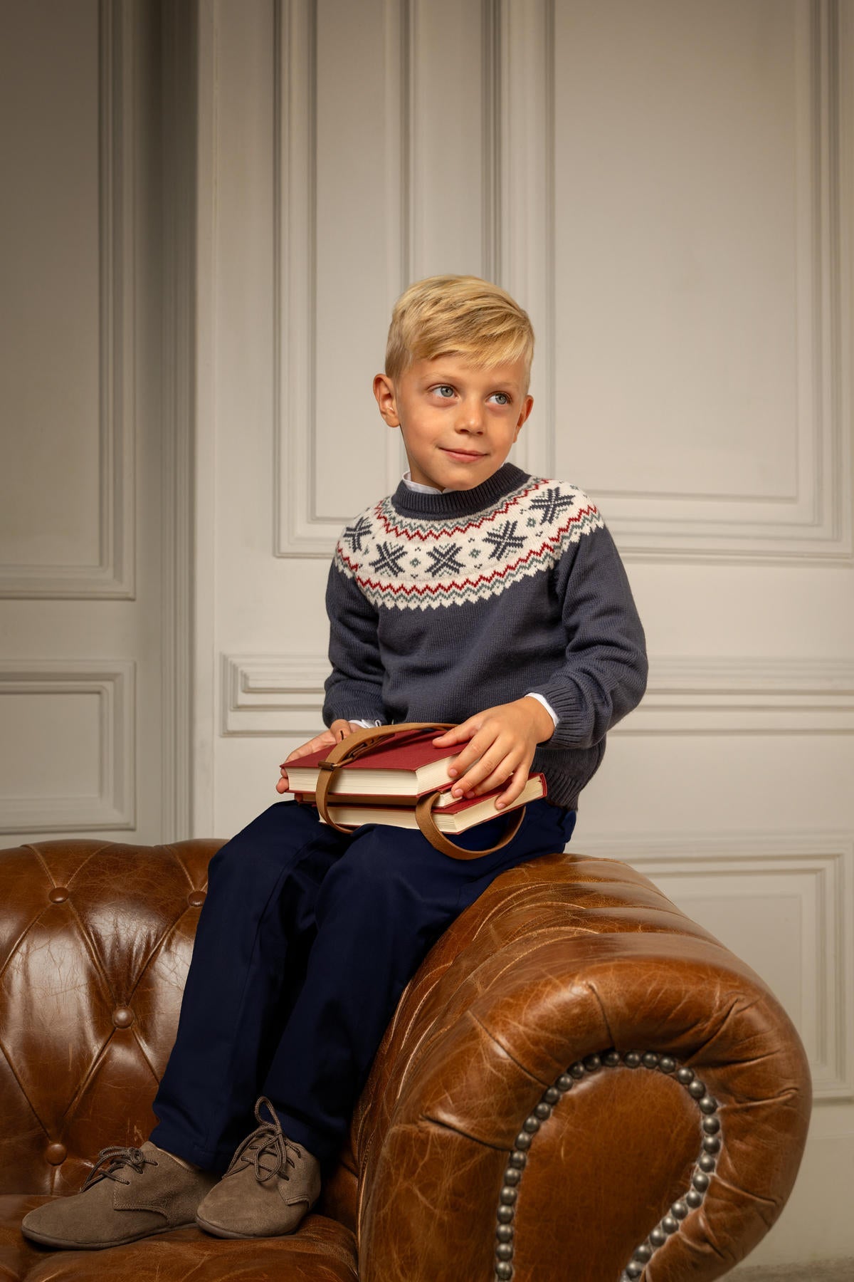 Boy seated on a leather sofa wearing deep navy George pants, holding stacked books against a paneled wall