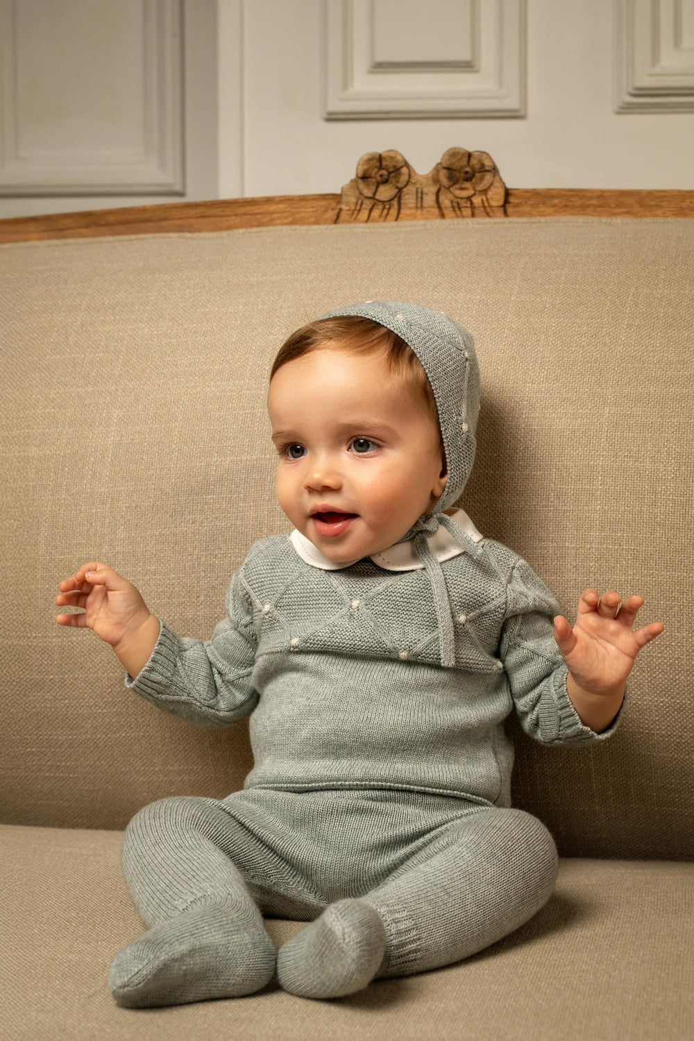 Front view of Dominique baby set in fern green with diamond knit sweater, matching bonnet and footed pants on a seated baby