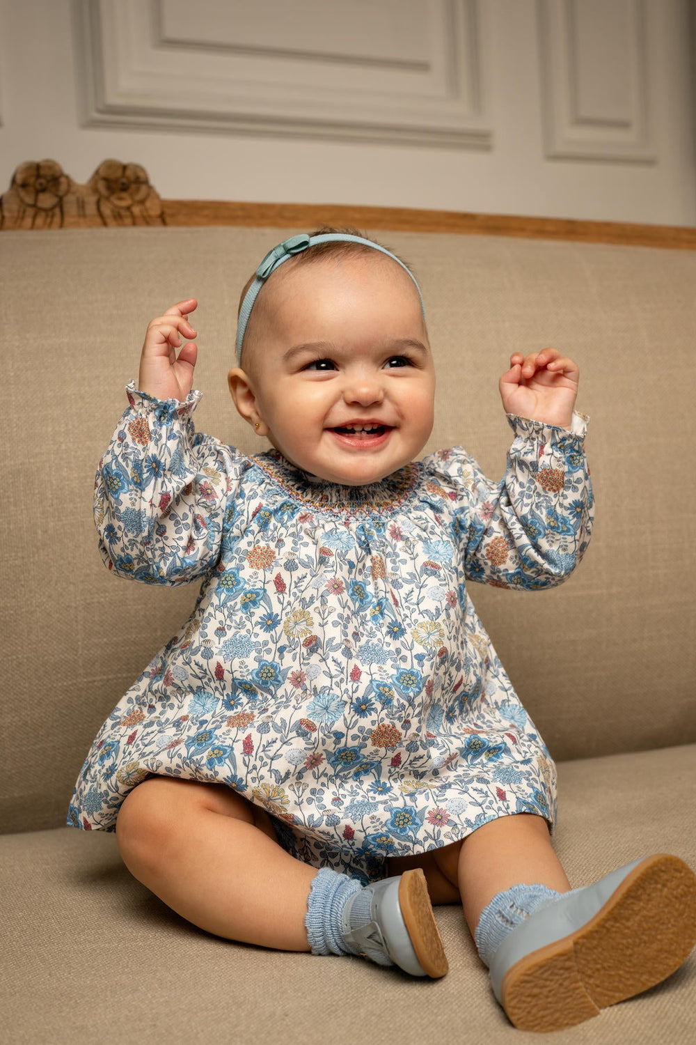 Front view of Cynthia short dress in Bluebell Garden showcasing smocked yoke, long puff sleeves, and floral print on a baby seated on a beige sofa