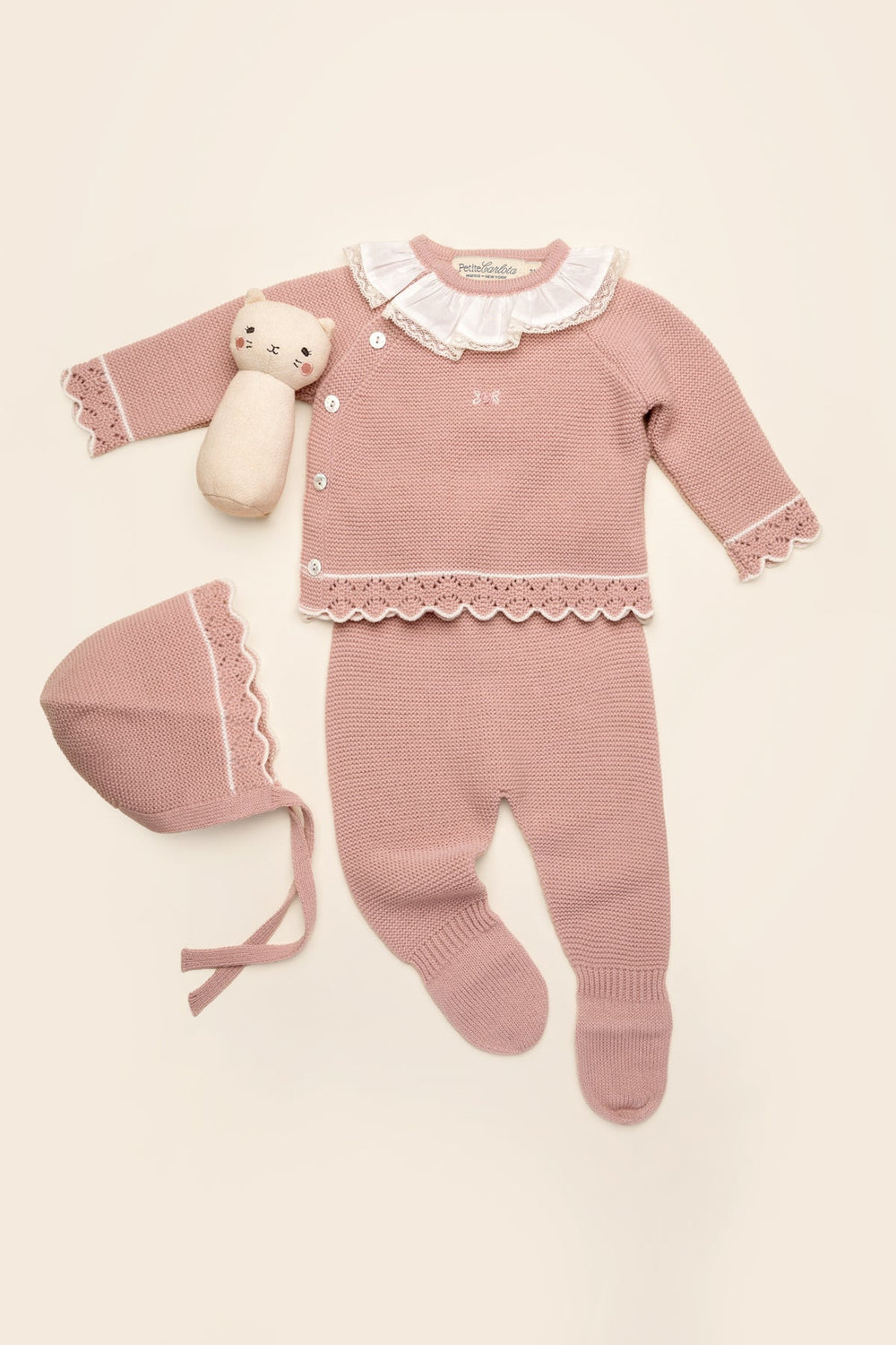 Flat lay of Bella baby set in rose petal with scalloped trim, ruffle collar, bonnet and footed pants on cream background