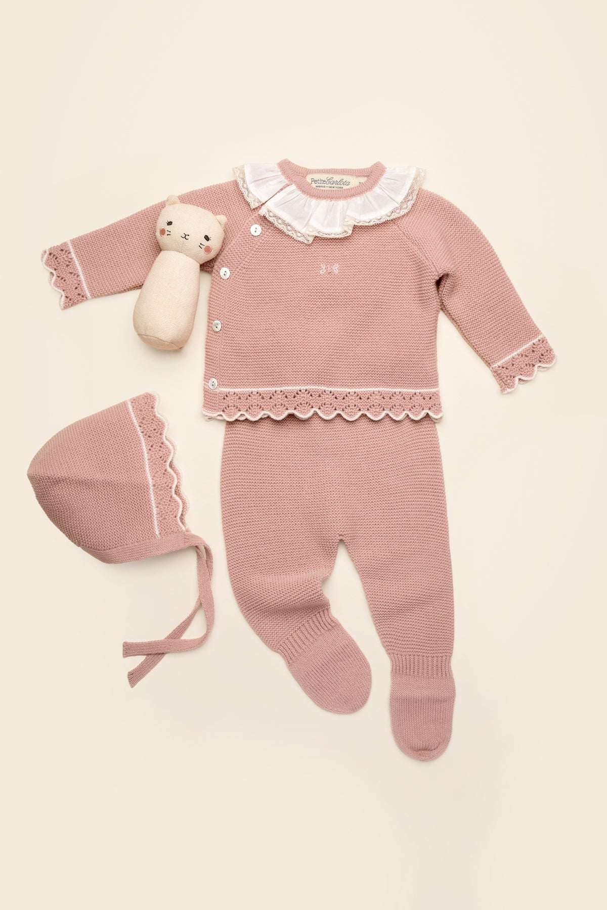 Flat lay of Bella baby set in rose petal with scalloped trim, ruffle collar, bonnet and footed pants on cream background