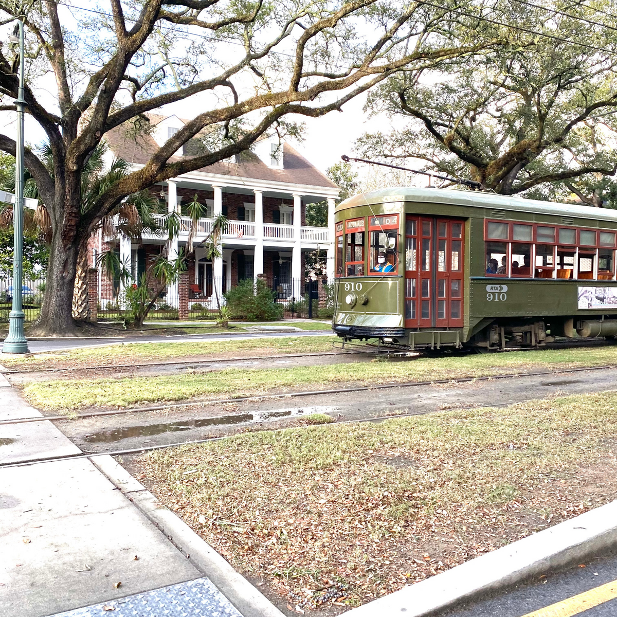 Green New Orleans streetcar 910 traveling past a two-story white-pillared house beneath large live oak trees
