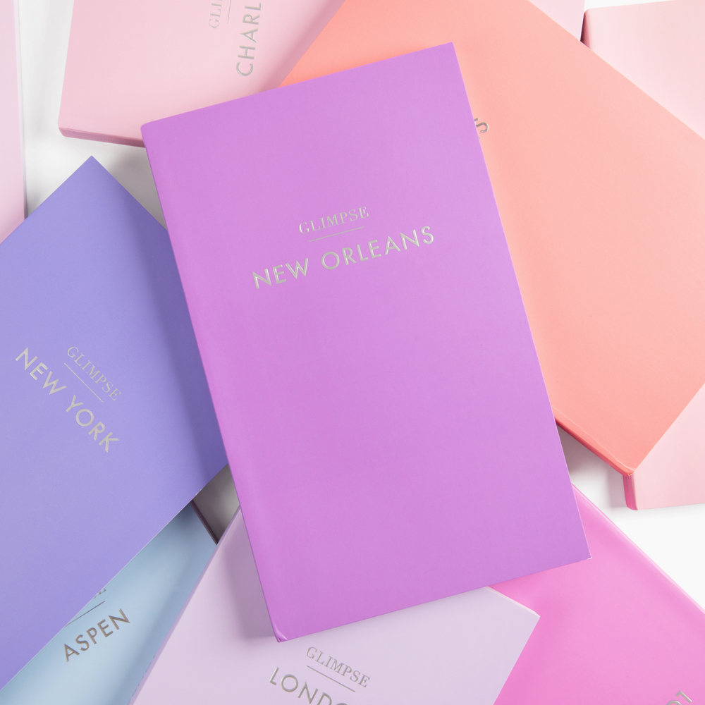 Top-down view of Glimpse Guide travel journals showcasing a purple NEW ORLEANS cover with gold foil title and pastel covers