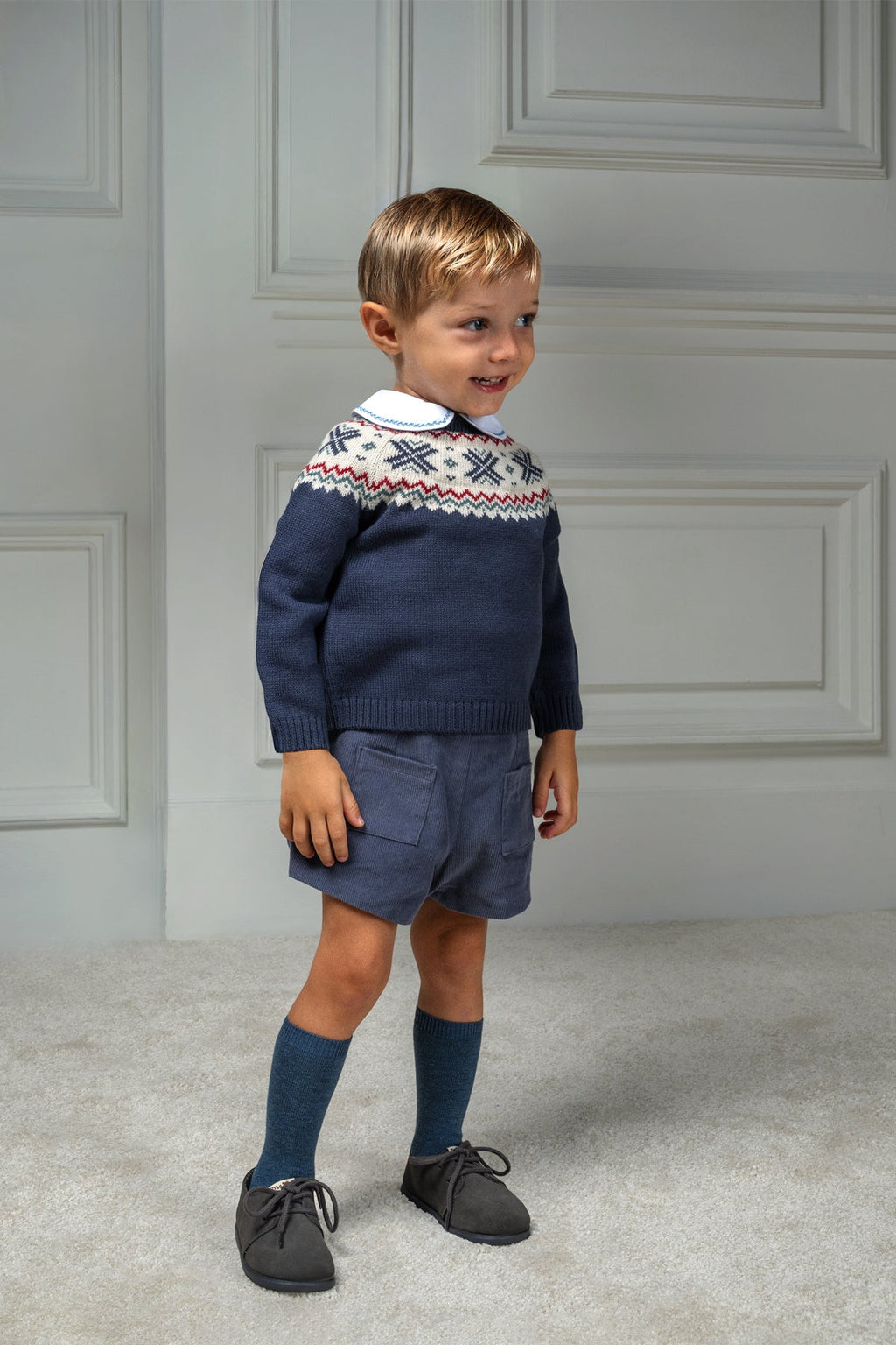 Child wearing a navy patterned sweater, shorts, and knee-high socks indoors