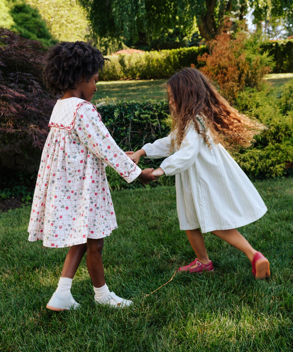 Two girls holding hands and spinning on a sunlit lawn, one wearing the Gigi Dress in Pomegranate Parade