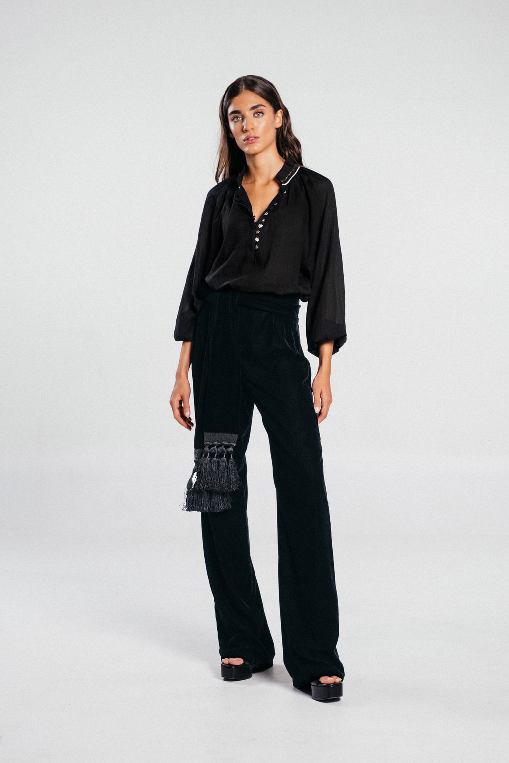 Full-length view of Mahtab straight-leg velvet pants in black, high waist with tassel wrap belt, light gray background