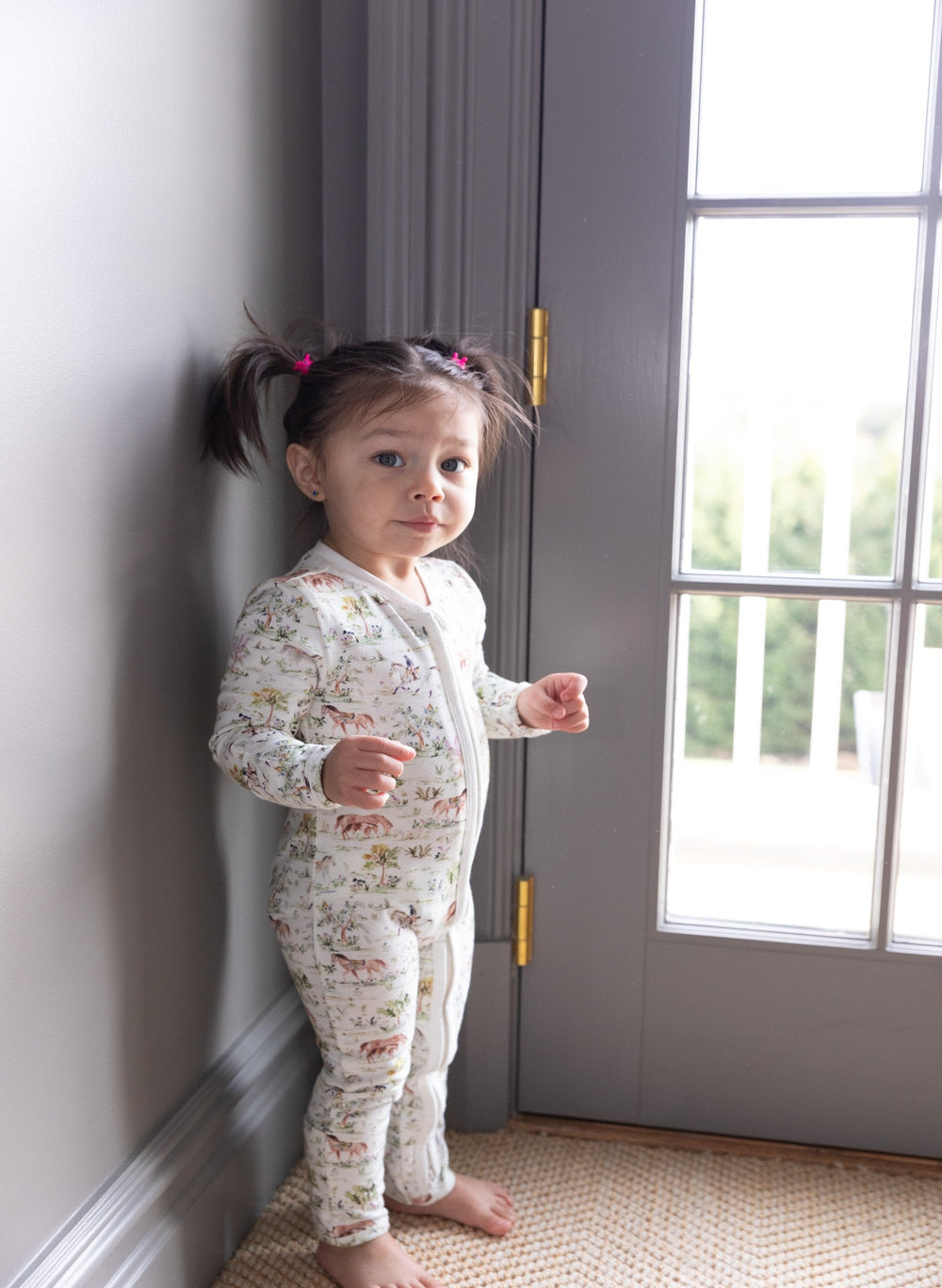 Full-length front view of Hunter Pace Horse One-Piece on a toddler showcasing horse print and front zipper by a gray door