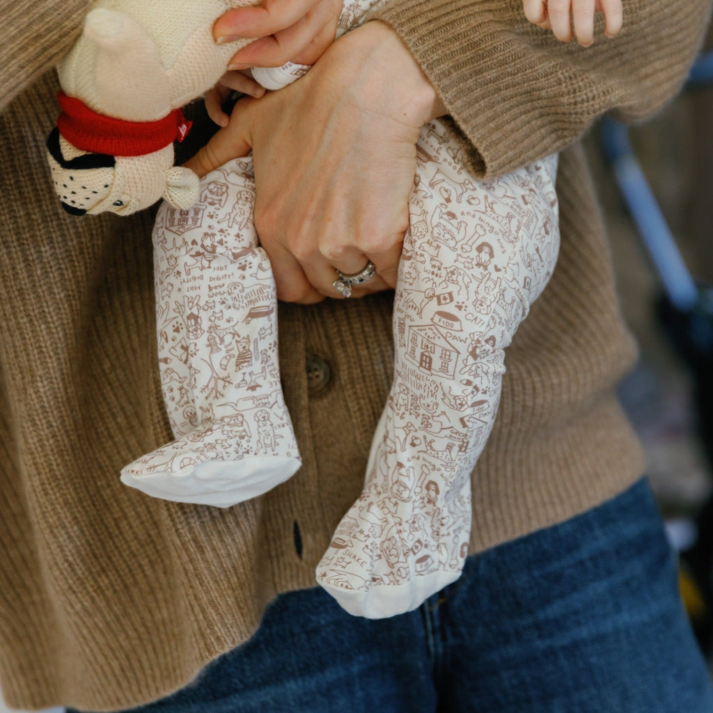 Close-up of Good Dog Zip Baby Onesie showcasing ivory dog print and brown striped footed trim on a baby