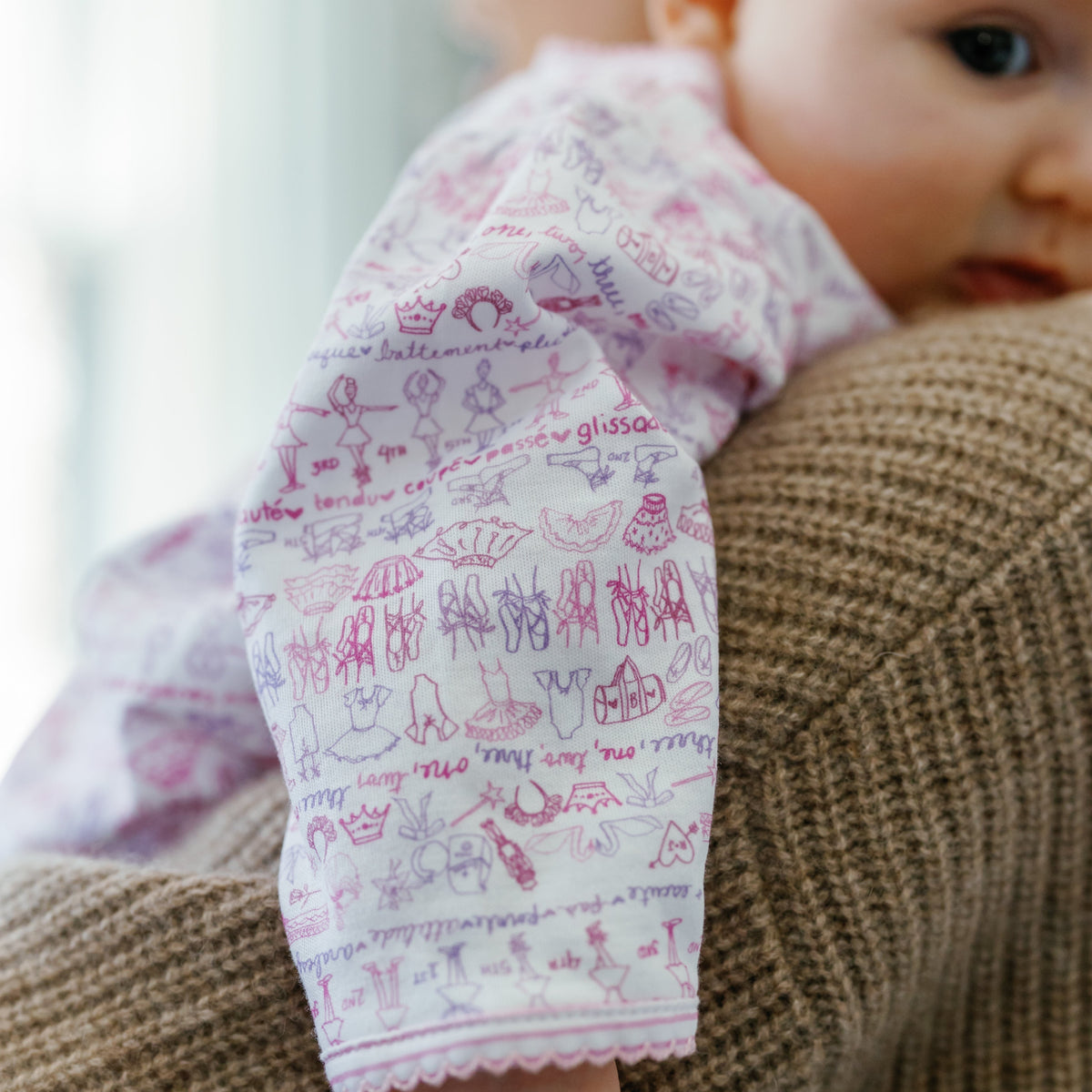 Side view of pink Ballet Snap baby onesie showcasing a ballet-themed print and scalloped sleeve against a knit background