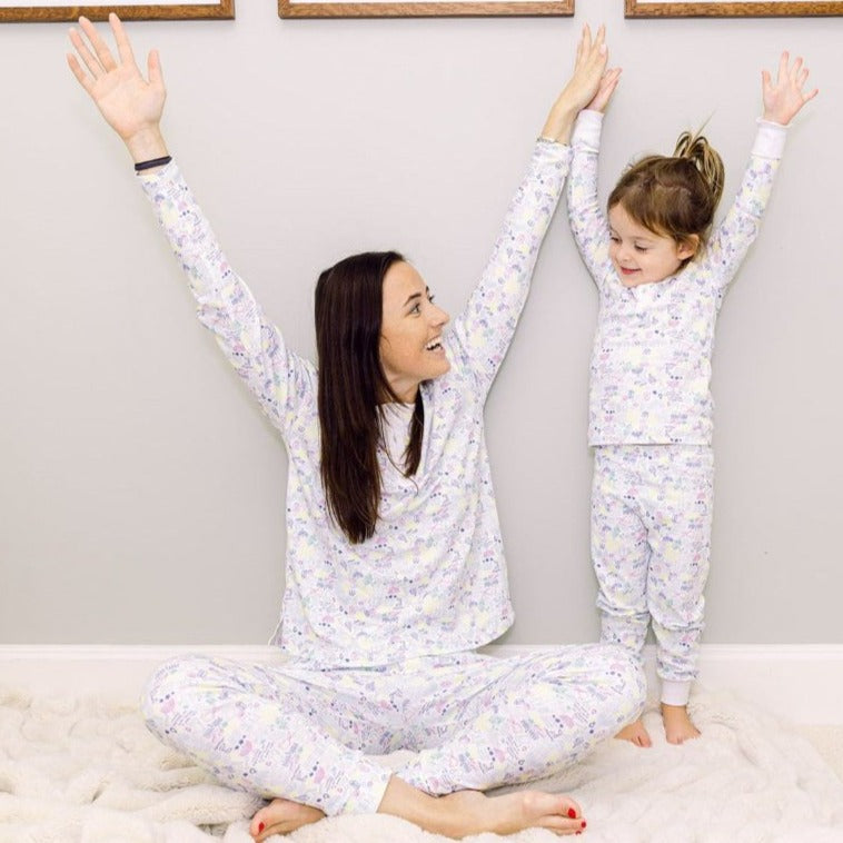 Mother and young daughter in matching Hanukkah-print pajamas raising their arms, smiling against a pale wall