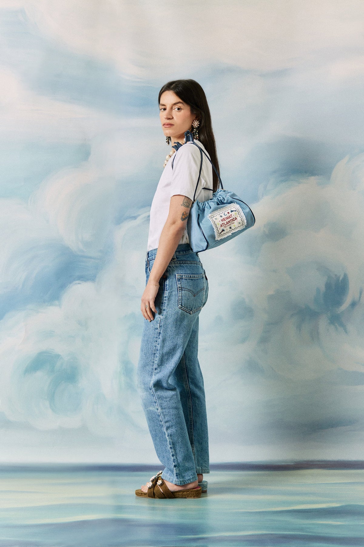 Model in profile wearing a blue striped drawstring pouch with HEIMAT ATLANTICA patch against a cloud-painted backdrop