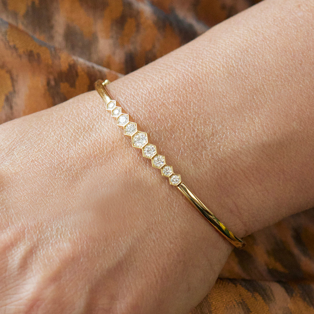 The Graduated Hexagon Bezel Set Diamond Bangle, showcasing alternating hexagon and round diamonds, sparkles on a wrist against a brown patterned fabric background.