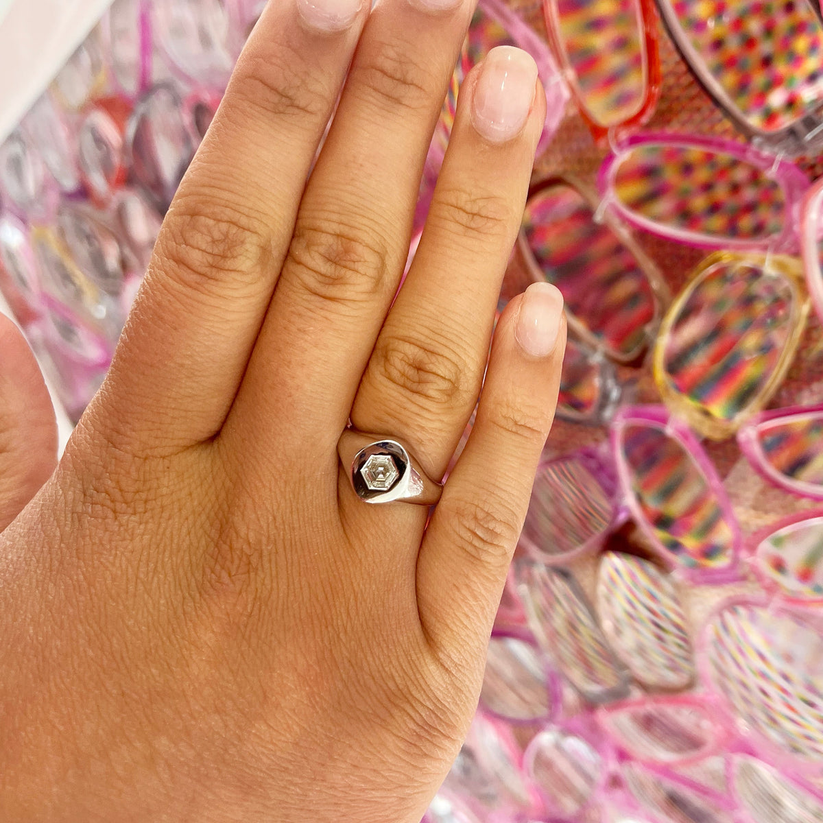 A hand with neatly manicured nails wears the Signet Ring with Burnish Set Hexagon Diamond, while colorful, patterned sunglasses are arranged in rows in the background.