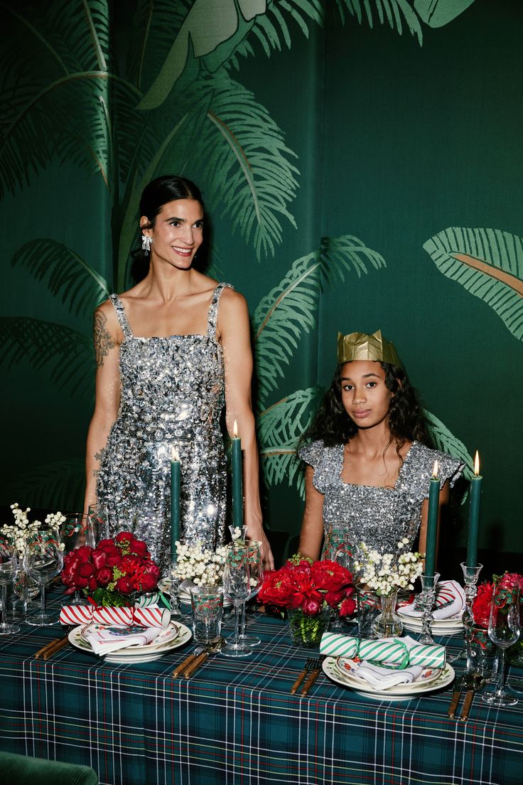 Two women at a candlelit holiday table with a green tartan tablecloth, red floral centerpieces, glassware, and a dark green botanical backdrop