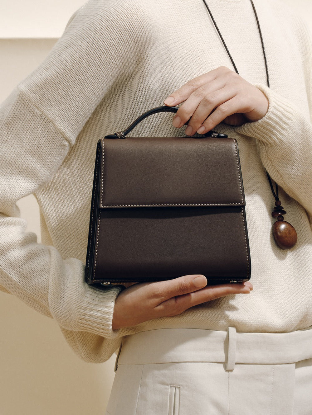 Front of The Small Top Handle handbag in dark brown nappa showing top handle and contrast stitching against cream sweater