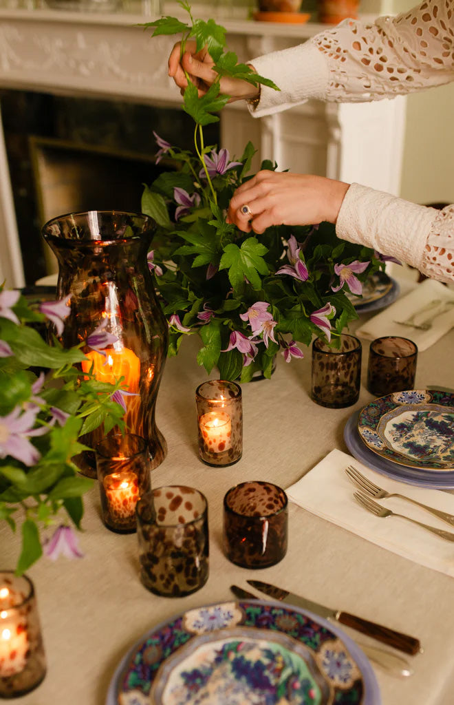 Amethyst marble hurricane vase glowing on a dining table with lit spotted glass votives while a person arranges purple flowers