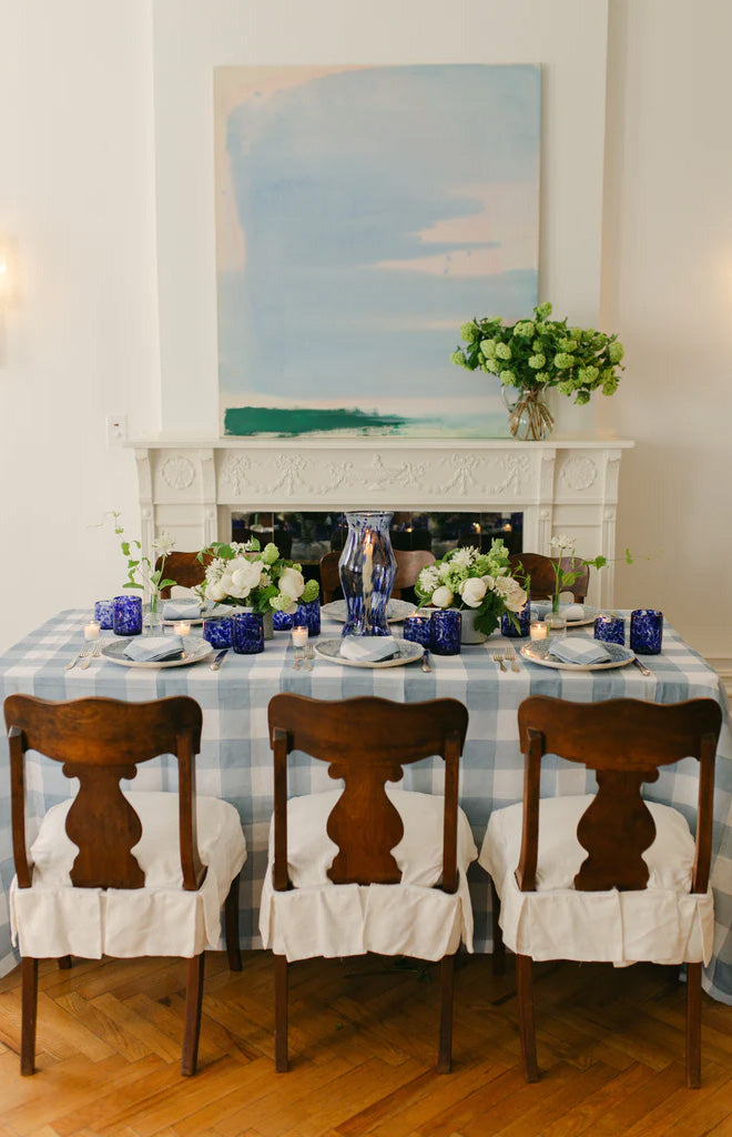 Blue marble hurricane vase as dining table centerpiece on blue check tablecloth with white flowers, wooden chairs, and fireplace