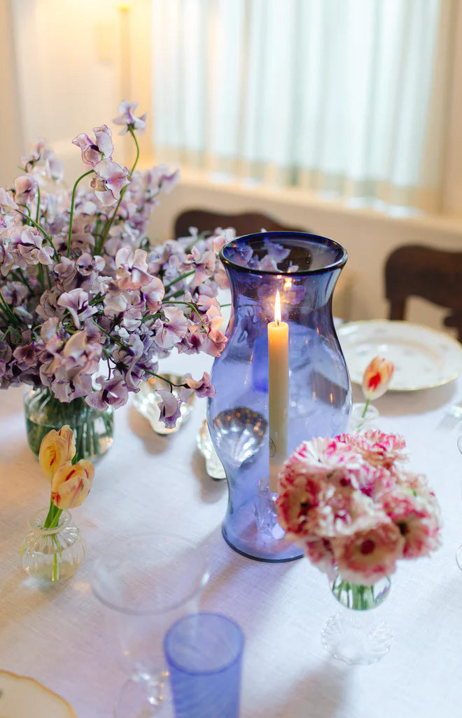 Front view of Hurricane vase in French blue showcasing bulbous hand‑blown glass holding a lit taper candle on a table with flowers