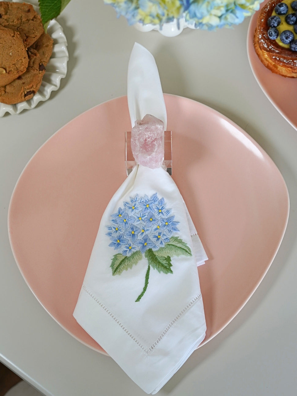 Top-down view of French Hydrangea dinner napkin showcasing hand-embroidered blue hydrangea and hemstitched trim on pink plate
