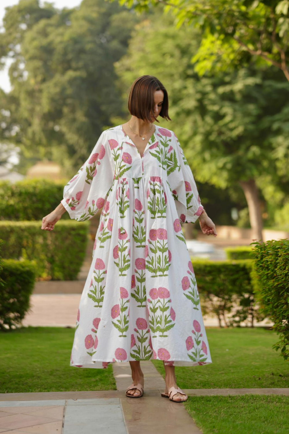 Front view of Taj Maxi Dress in pink flower print showcasing puffed 3/4 sleeves, gathered waist and flowy fit on model in a garden