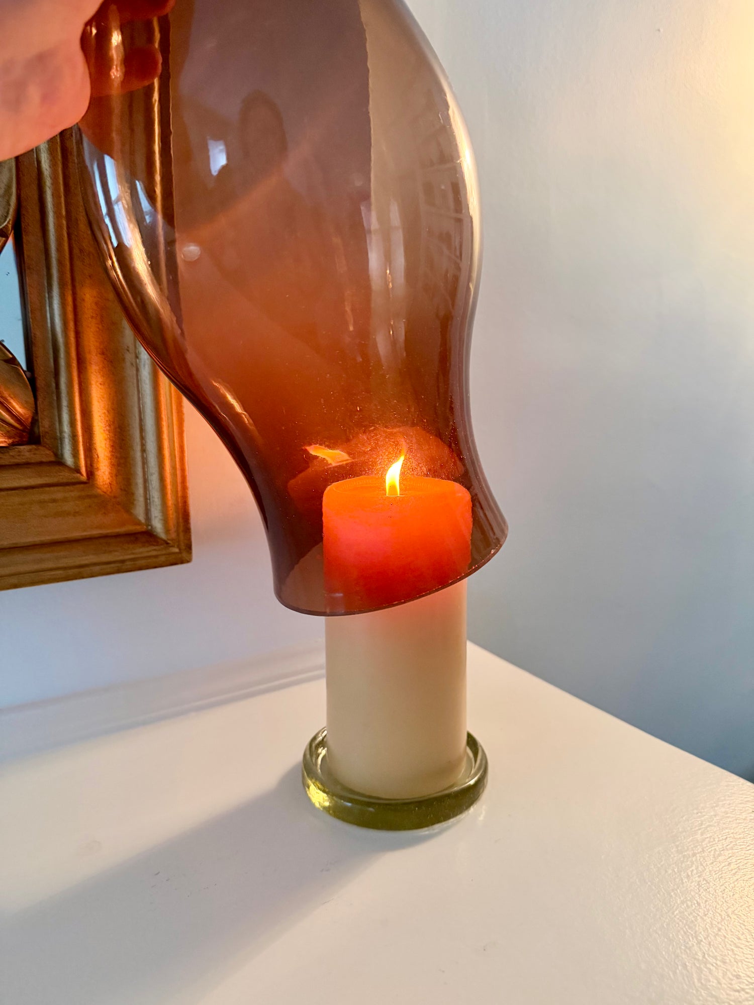 Close-up of clear glass trivet under a lit pillar candle, shown fitting inside a brown glass hurricane on a white tabletop
