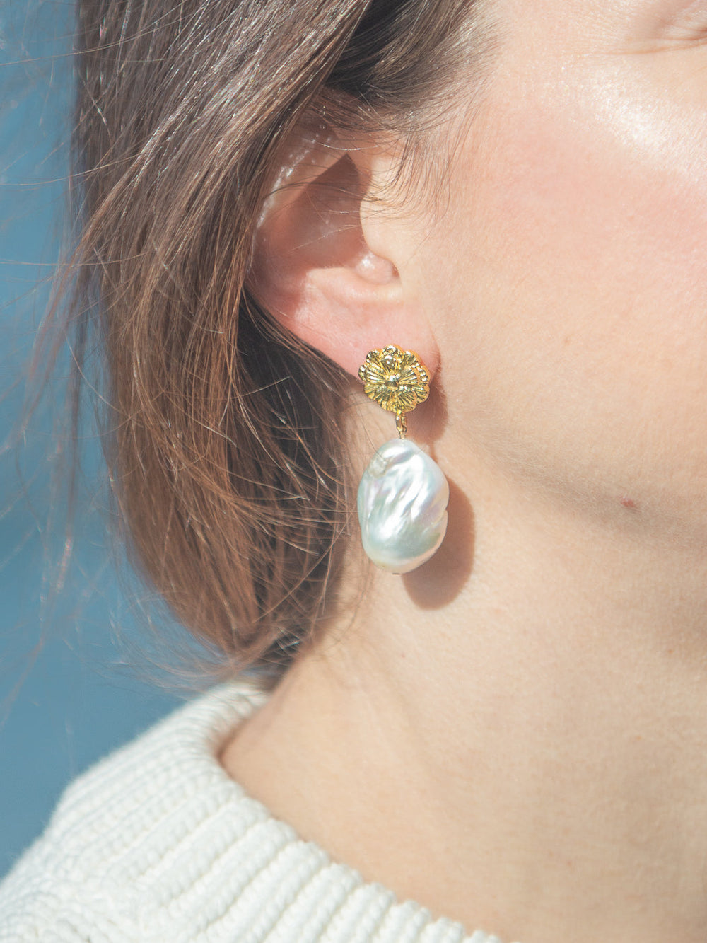 Close-up of Mindy earrings showing gold-plated floral stud and dangling freshwater baroque pearl on blurred blue background