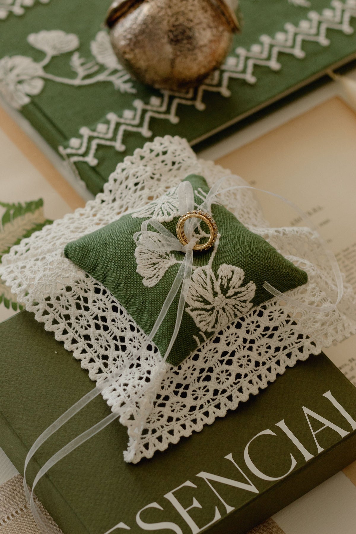 Top down view of Cosmo Lace pillow in olive with white crochet lace trim, organza ribbon and two gold rings on a green box