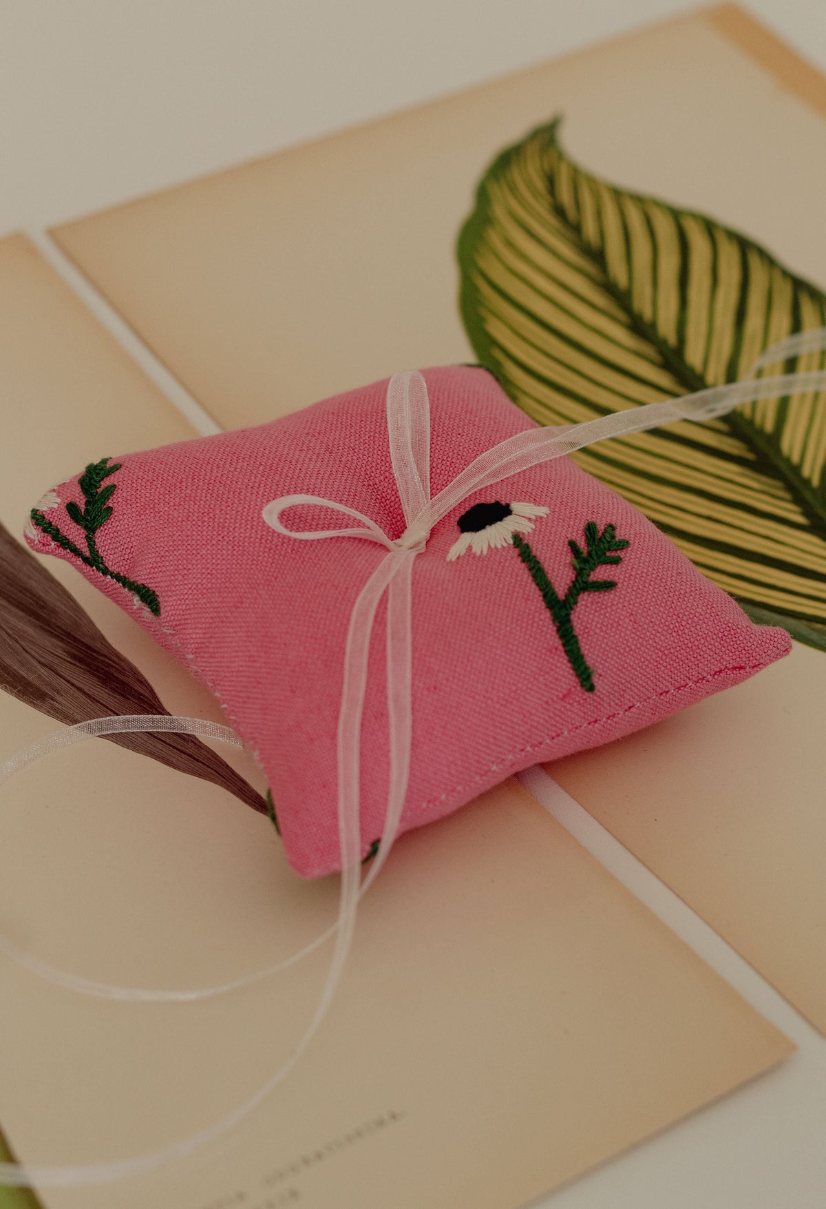 Angled top view of Manzanilla ring pillow showcasing pink linen fabric, embroidered white daisy motifs, and tied organza ribbon on a cream paper background
