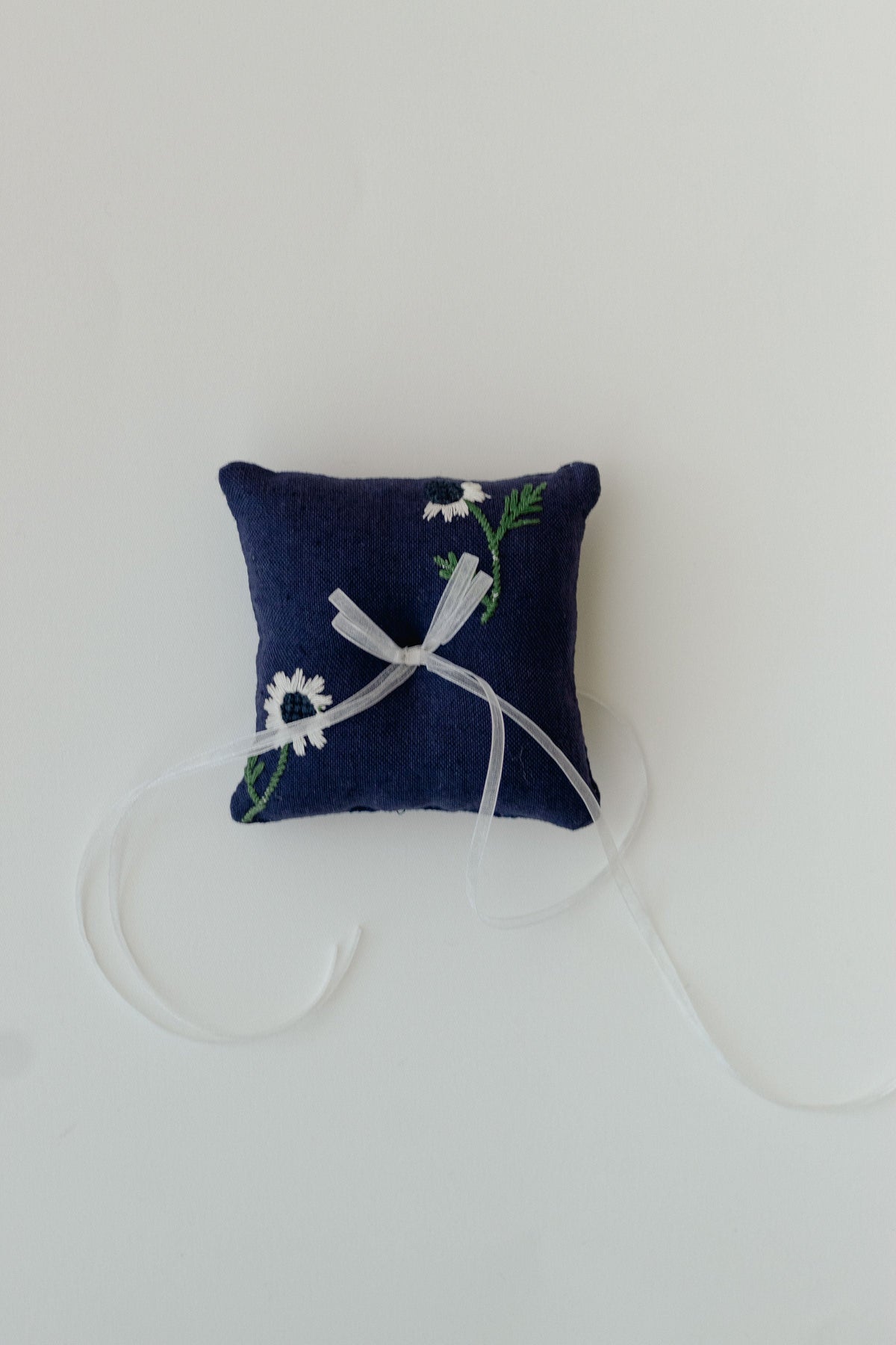Top view of Manzanilla ring pillow in navy showcasing white daisy embroidery and organza ribbon on white background