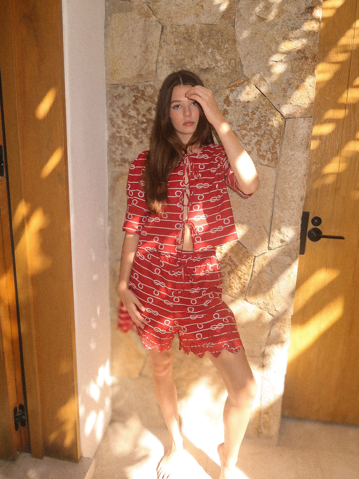 Front view of model wearing red scalloped tie-front top and matching shorts in anchor rope print, sunlit stone wall background