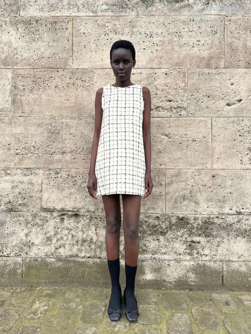 Full-length front view of Jane Dress in Carré Alpin showing sleeveless white checked tweed shift with raw-edge hem on a model against a stone wall