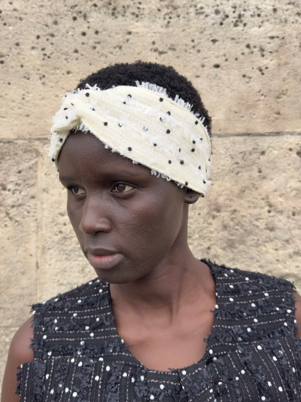 Three-quarter view of Petit Pois tweed headband on model showing twisted front knot, black dot texture and elastic back, stone wall background