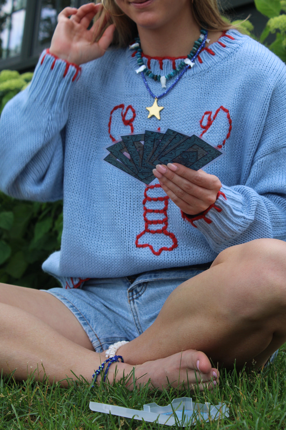 Model seated on grass holding cards, wearing Scaly Blue Fish necklace with blue chip beads and mother-of-pearl fish