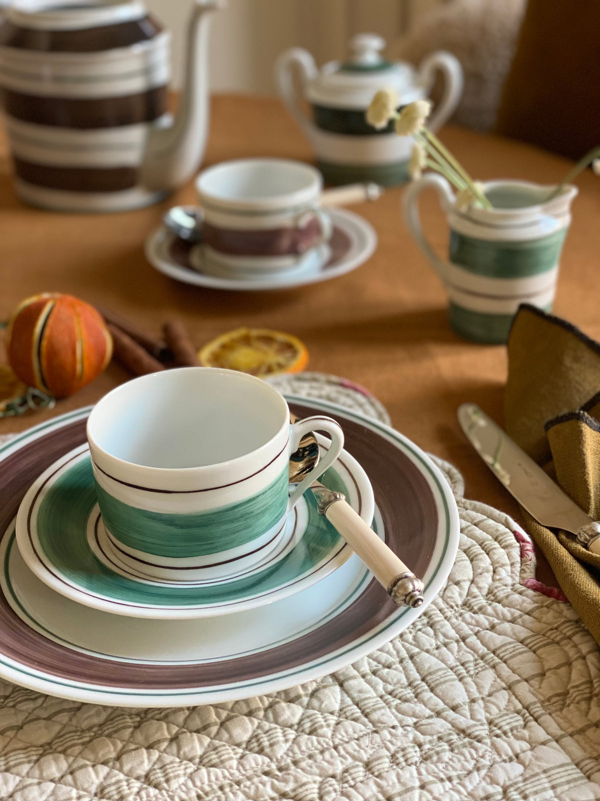 Close-up of Tangerine tea cup with plate showing hand-painted green brushstroke band and brown rim on quilted tablecloth