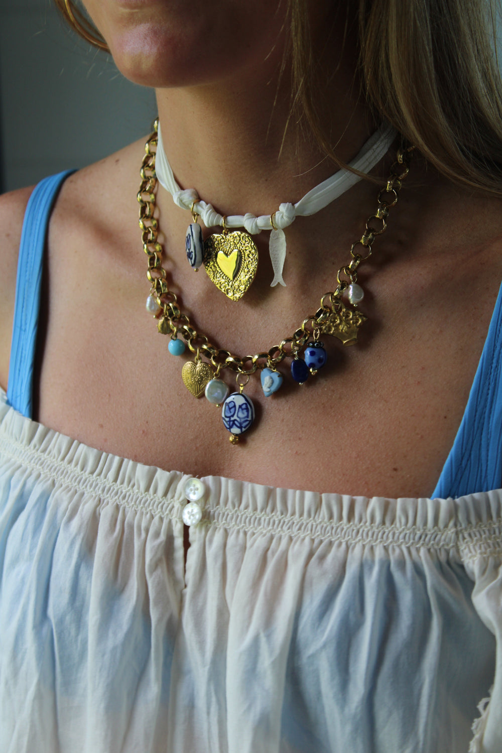 Close-up of White Ribbon Charm Necklace on a model, showcasing white silk ribbon with clear fish, blue tulip, and gold floral heart charms against a blue tank and white top
