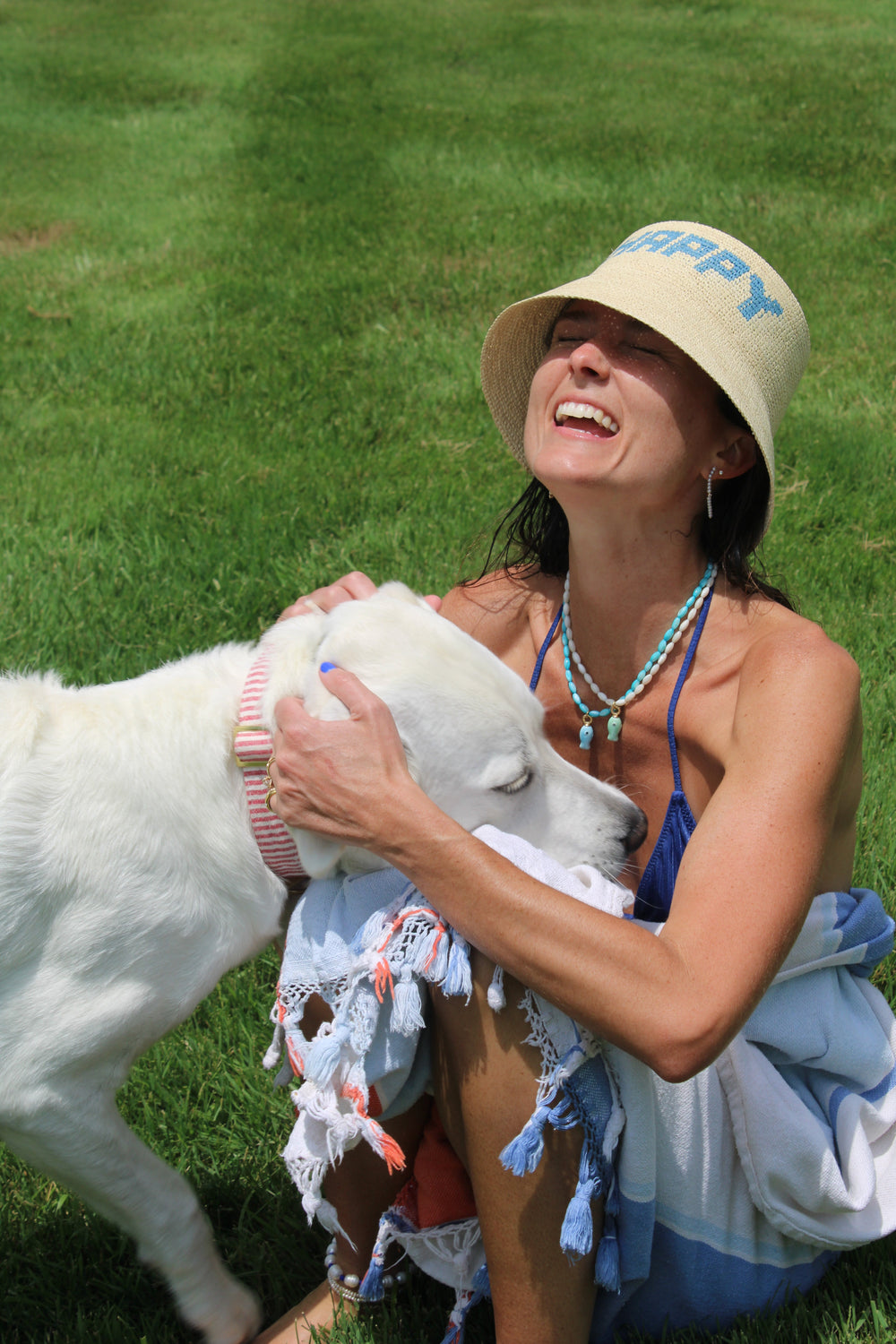 Woman smiling and petting a white dog while wearing a light blue beaded fish necklace on a grassy lawn