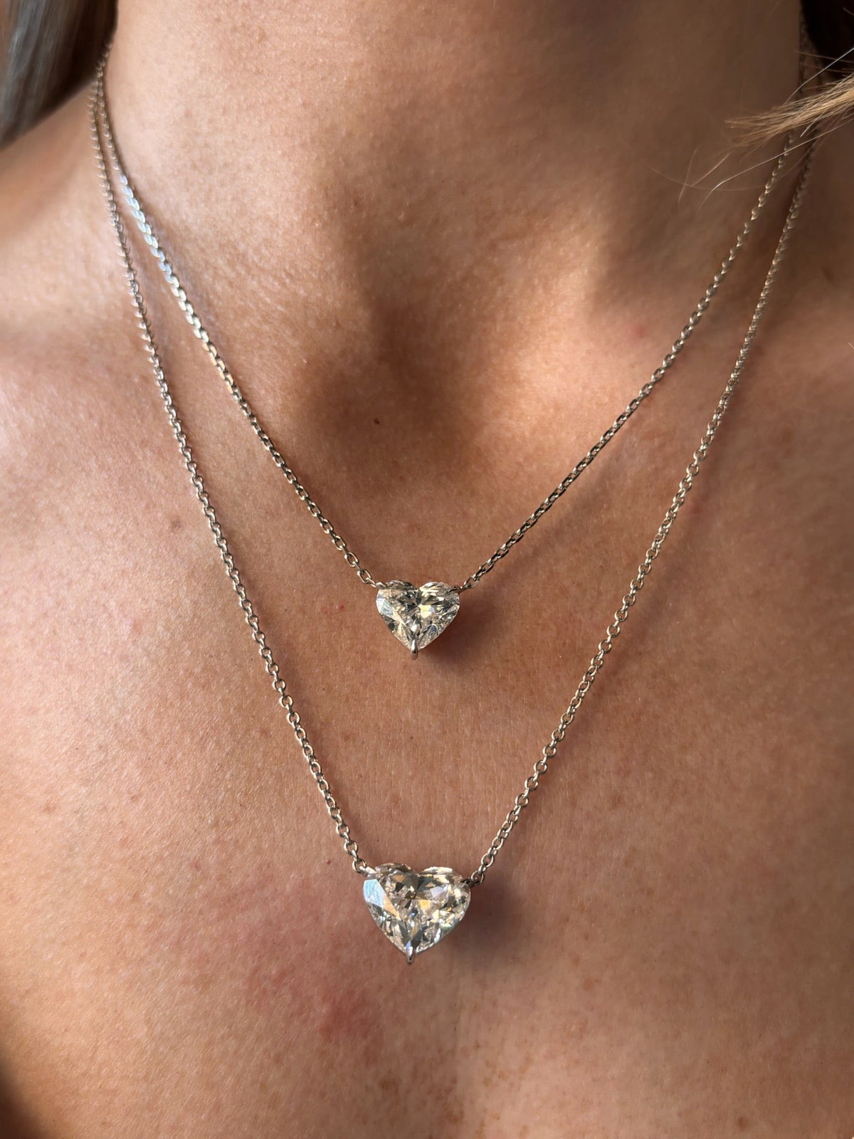 A close-up of a person’s neck wearing two layered Heart-Shaped Diamond Solitaire Pendant necklaces, each featuring a sparkling heart-shaped diamond, with one pendant resting above the other.