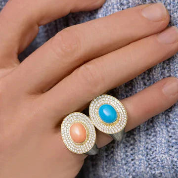 Hand wearing two Eyecon rings with turquoise and coral oval cabochons and pave diamond halos on 18k yellow gold