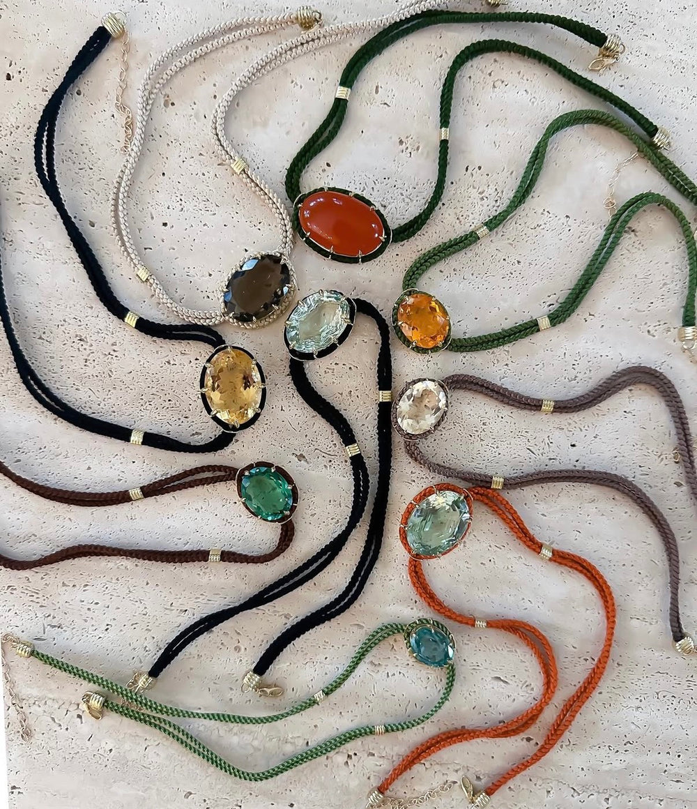 Flat lay of silk cord pendants with assorted colored gemstones set in 18K yellow gold on a textured stone background
