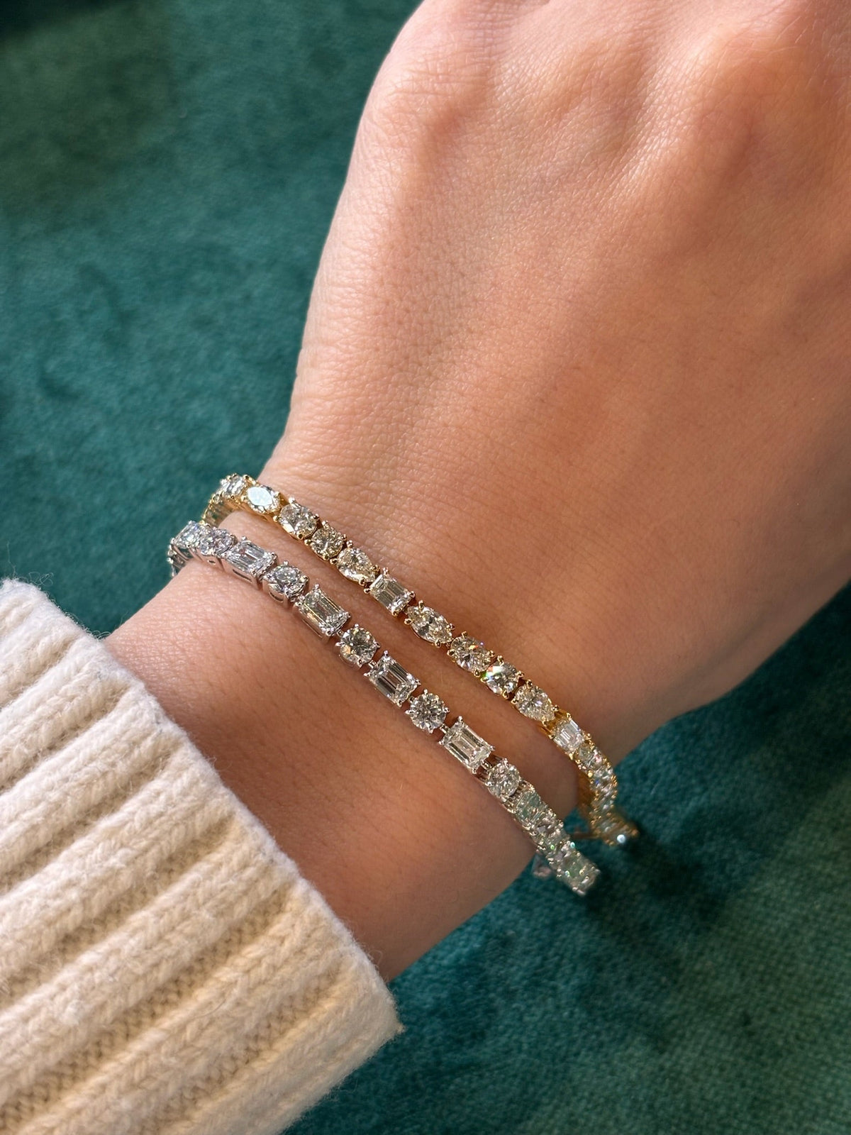 A hand in a cream-colored knit sweater displays the Alternating East-West Set Round and Emerald Cut Diamond Tennis Bracelet in White Gold against a green background.