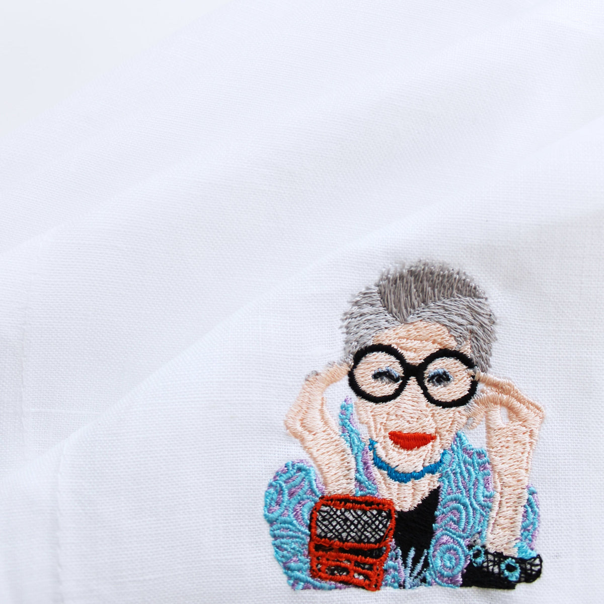 Close-up of cocktail napkin showing embroidered Iris Apfel portrait with black round glasses and turquoise jacket on white linen