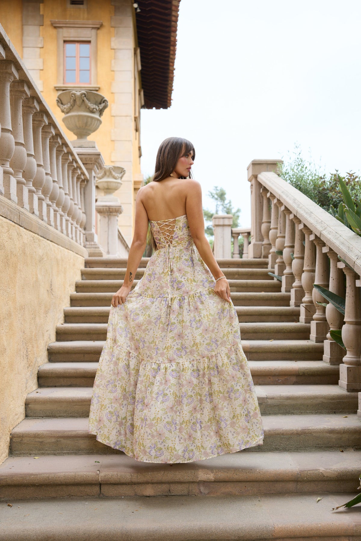 Rear view of The Isabel Dress in Cordoba Print showcasing floral organza, corset lacing, and tiered skirt on stone stairs