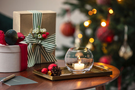 Clear glass tealight holder burning a tealight on a tray with wrapped gifts and a decorated Christmas tree in the background