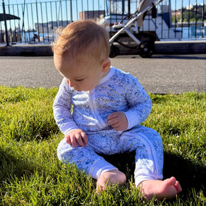 Front view of the blue Tennis Zip Baby Onesie showcasing its hand-drawn tennis print on a seated baby outdoors